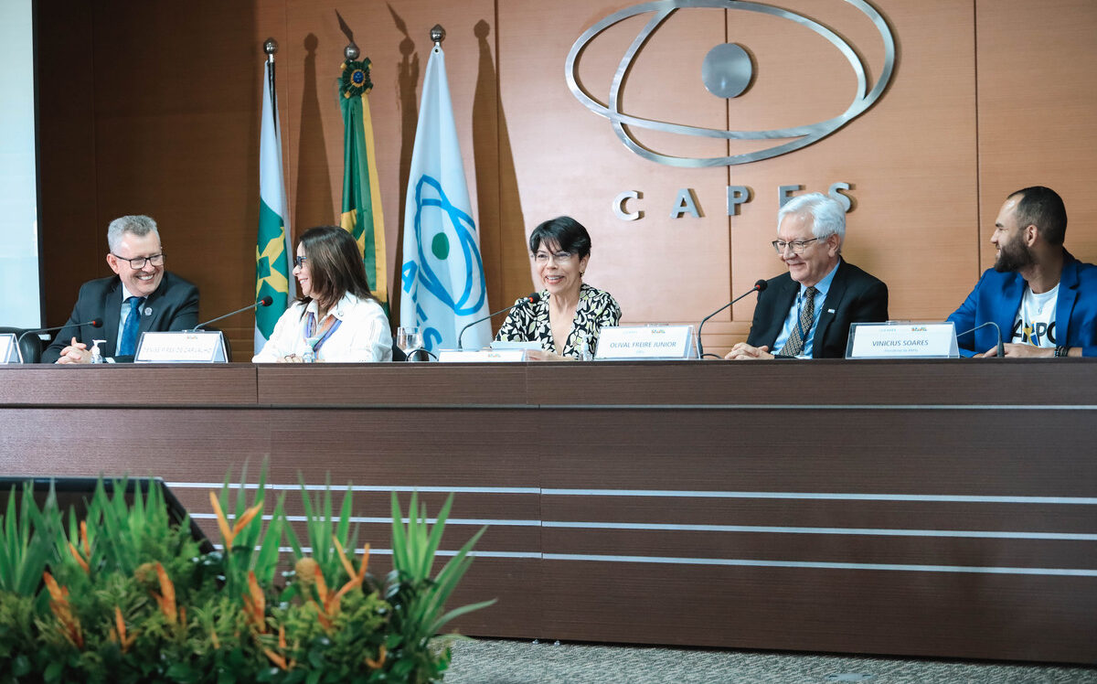 “Cerimônia do Prêmio CAPES de Tese na sede da CAPES, em Brasília. Na mesa diretora, cinco participantes estão sentados frente ao público, com placas de identificação. Ao fundo, aparecem as bandeiras do Brasil, do MEC e da CAPES, além do logotipo metálico da agência na parede. Os integrantes da mesa conversam e sorriem durante o evento, em um ambiente formal e iluminado.