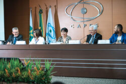 “Cerimônia do Prêmio CAPES de Tese na sede da CAPES, em Brasília. Na mesa diretora, cinco participantes estão sentados frente ao público, com placas de identificação. Ao fundo, aparecem as bandeiras do Brasil, do MEC e da CAPES, além do logotipo metálico da agência na parede. Os integrantes da mesa conversam e sorriem durante o evento, em um ambiente formal e iluminado.