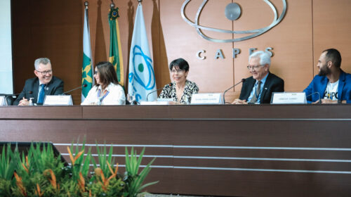 “Cerimônia do Prêmio CAPES de Tese na sede da CAPES, em Brasília. Na mesa diretora, cinco participantes estão sentados frente ao público, com placas de identificação. Ao fundo, aparecem as bandeiras do Brasil, do MEC e da CAPES, além do logotipo metálico da agência na parede. Os integrantes da mesa conversam e sorriem durante o evento, em um ambiente formal e iluminado.
