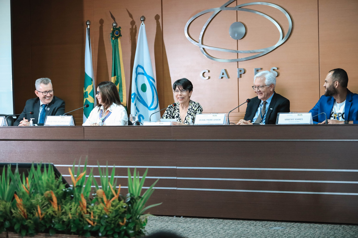 “Cerimônia do Prêmio CAPES de Tese na sede da CAPES, em Brasília. Na mesa diretora, cinco participantes estão sentados frente ao público, com placas de identificação. Ao fundo, aparecem as bandeiras do Brasil, do MEC e da CAPES, além do logotipo metálico da agência na parede. Os integrantes da mesa conversam e sorriem durante o evento, em um ambiente formal e iluminado.