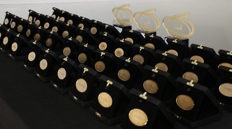 Várias medalhas do Prêmio CAPES de Tese expostas em estojos pretos sobre uma mesa, organizadas em fileiras. Ao fundo, três troféus transparentes com o símbolo da CAPES.