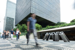 Pessoas caminham em frente ao edifício moderno do BNDES, no centro do Rio de Janeiro. A fachada de vidro do prédio se destaca ao fundo, enquanto o letreiro “BNDES” aparece em primeiro plano. Algumas pessoas estão em movimento, levemente desfocadas, indicando circulação urbana.