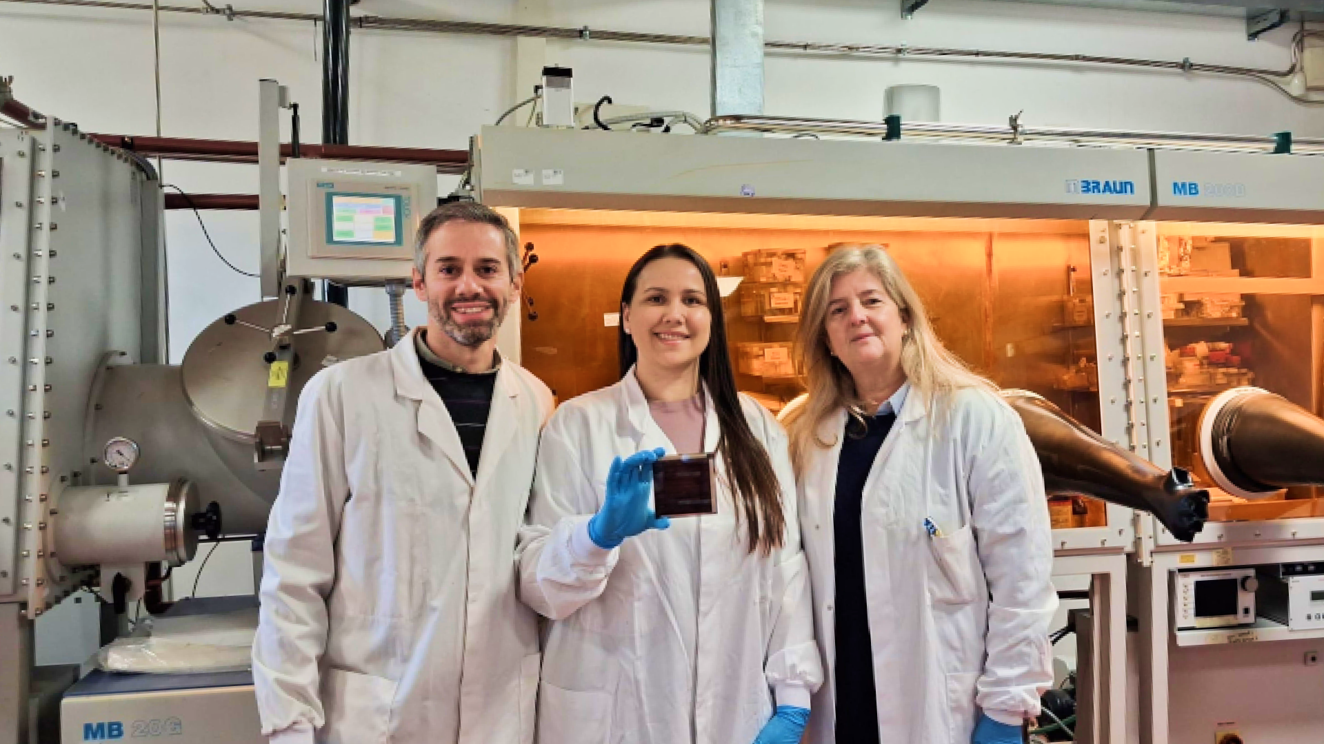 Três pesquisadores usando jalecos e luvas, posam em laboratório de pesquisa em energia solar. Ao centro, uma pesquisadora segura um dispositivo de célula solar de perovskita, com equipamentos científicos ao fundo.