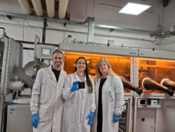 Três pesquisadores, usando jalecos e luvas, posam em laboratório de pesquisa em energia solar. Ao centro, uma pesquisadora segura um dispositivo de célula solar de perovskita, com equipamentos científicos ao fundo.