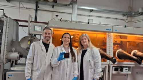 Três pesquisadores, usando jalecos e luvas, posam em laboratório de pesquisa em energia solar. Ao centro, uma pesquisadora segura um dispositivo de célula solar de perovskita, com equipamentos científicos ao fundo.