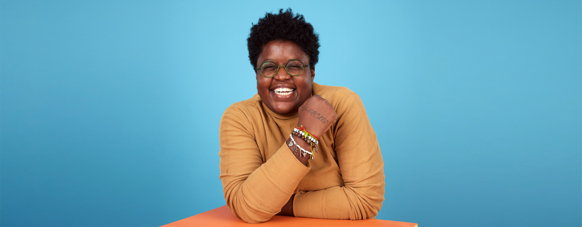 Retrato de uma mulher negra jovem sorridente, com cabelo afro curto e volumoso, usando óculos verdes de armação redonda. Ela veste um suéter bege de gola alta, exibe pulseiras coloridas no pulso direito e está sentada com os braços apoiados em uma mesa laranja, sobre fundo azul claro.